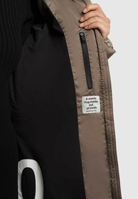 Hand holds open taupe jacket showing black inner lining with a zipper pocket and white label reading "A warm hug made out of cloth."
