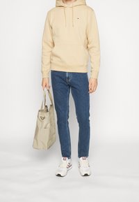Sudadera con capucha beige, vaqueros azules y zapatillas blancas con detalles en rosa y gris; lleva un bolso tote color beige con apertura superior y bolsillo lateral.