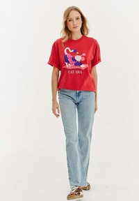 Czerwona koszulka z krótkim rękawem z zabawnym wzorem kota oraz napisem "ERA KOTÓW", w połączeniu z jasnoniebieskimi dżinsami i trampkami w wzór zwierzęcy.
