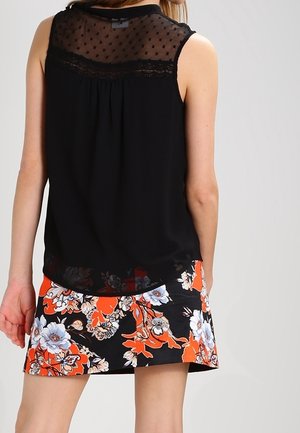 Kobieta nosząca czarny top bez rękawów z przezroczystą górą w groszki oraz czarną spódnicę w kwiaty z jaskrawymi pomarańczowymi i białymi kwiatami.