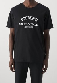 Mustassa lyhyessä hihassa puuvillapaidassa on valkoiset tekstit "ICEBERG MILANO ITALIA since 1974", jotka on taitettu kaarevaksi muotoon rinnan kohdalla.