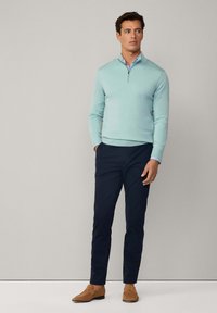 Helleblaue Strickjacke mit Reißverschlusskragen, kombiniert mit dunkelblauen Hosen und braunen Slippern. Glatte Textur und taillierte Passform. Minimalistisches Design.