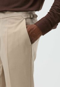 Beige, op maat gemaakte broek met zijzakken, voorzien van een riemgespaccent bij de taille. De stof heeft een gladde textuur.