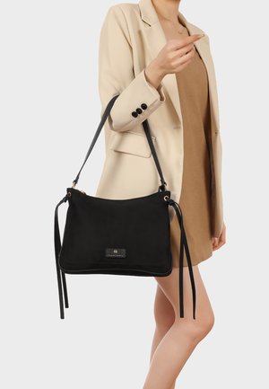 Donna con blazer beige e vestito marrone che tiene una borsa nera a tracolla con spalline lunghe e toppa con logo frontale.
