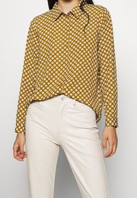 Camicia gialla senape con bottoni, caratterizzata da motivi floreali bianchi, maniche lunghe e colletto classico, abbinata a pantaloni color crema.