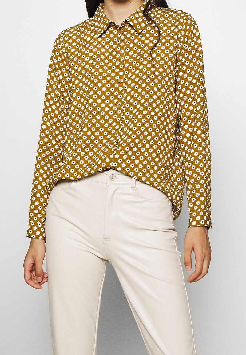 Camicia gialla senape con bottoni, caratterizzata da motivi floreali bianchi, maniche lunghe e colletto classico, abbinata a pantaloni color crema.
