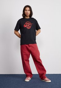 Juodas medvilnės marškinėliai su raudonuoju gyvate ir logotipu priekyje, derinami su laisvomis raudonomis džinsinėmis kelnėmis ir rožiniais sportbačiais.