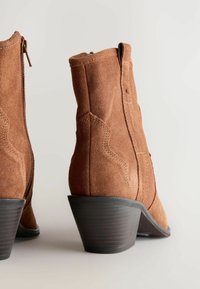 Botas de tobillo de ante marrón con un tacón apilado y cónico, y cierre de cremallera. Presentan costuras dobles y un acabado suave en el tacón.