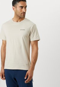T-shirt en coton beige à manches courtes, col rond et petit logo Columbia noir sur la poitrine gauche. Texture lisse.