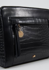 Schwarze Lederhandtasche mit Krokodilprägung, ausgestattet mit einer Reißverschlusstasche, einem Quasten-Detail und einem goldfarbenen Logo-Akzent.