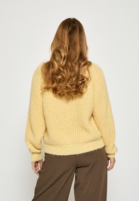 Dzeltena adīta džemperis ar teksturētu apdari, kas piedāvā brīvu piegriezumu un lielas pietekas piedurknes, apvienots ar brūnām plašajām biksēm.