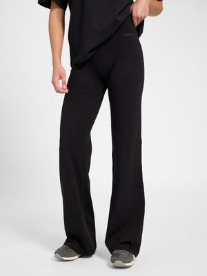 Persoon draagt zwarte uitlopende broek en zwart overhemd, staat met één hand bij de taille, grijze sneakers zichtbaar.