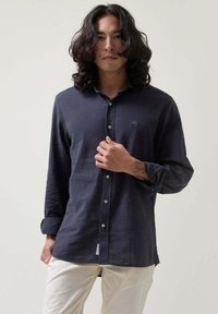 Camisa azul marino de lino con cuello, mangas largas y un pequeño logotipo bordado en el pecho. Combinada con pantalones claros.