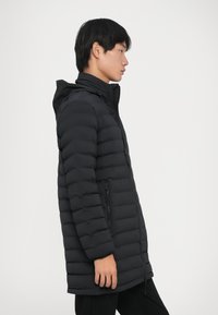 Giovane uomo in un cappotto invernale imbottito nero con cappuccio, in piedi di profilo a destra contro uno sfondo bianco uniforme.