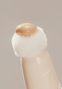 Aplikator do makijażu z beżowym kremowym końcem i białą puszystą główką, z gładką, zakrzywioną, jasnoróżową rączką.