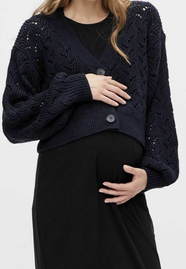 Gravid kvinna i en marinblå stickad cardigan och svart klänning, som varsamt omfamnar sin mage med båda händerna.