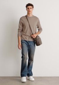 Pulover tricotat de culoare maro deschis, blugi albaștri și adidași albi; model purtând o geantă gri mică de umăr, stând pe un fundal neutru.