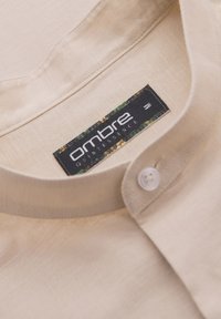 Col de chemise beige avec un bouton blanc et une étiquette noire affichant le nom de la marque Ombre Quintessence et la taille M.