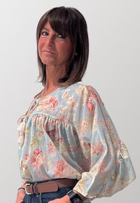 Blusa floreale in tessuto azzurro chiaro con maniche a palloncino, dettagli arricciati e motivi floreali in rosa e crema. Cintura marrone e jeans di denim scuro.