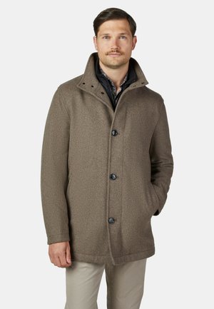 Mann trägt einen braunen wollenen Mantel mit Knöpfen über einer schwarzen gesteppte Jacke, mit einer Hand in der Tasche, steht vor einem schlichten hellen Hintergrund.