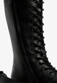 Black leather knee-high boot featuring a lace-up front, side zipper, and metal eyelets for laces. Smooth texture with a sleek finish.