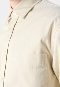 Chemise beige à boutons avec un col et une poche poitrine brodée du mot "BOSS" en fil blanc.
