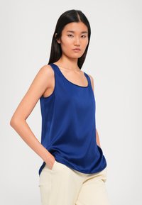 Young woman with long black hair wearing a sleeveless blue satin top and cream pants, hands in pockets, facing forward against white background.