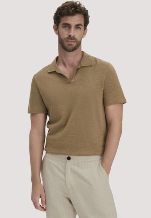 Mann mit lockigem braunem Haar und Bart, der ein braunes kurzärmeliges Poloshirt und hellbeige Hosen trägt, eine Hand in der Tasche, neutraler Hintergrund.