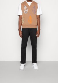 Beiger Strickwest mit V-Ausschnitt, verziert mit bunten Blattmustern in Blau, Orange, Lila und Grün, getragen über einem weißen T-Shirt und schwarzen Hosen.