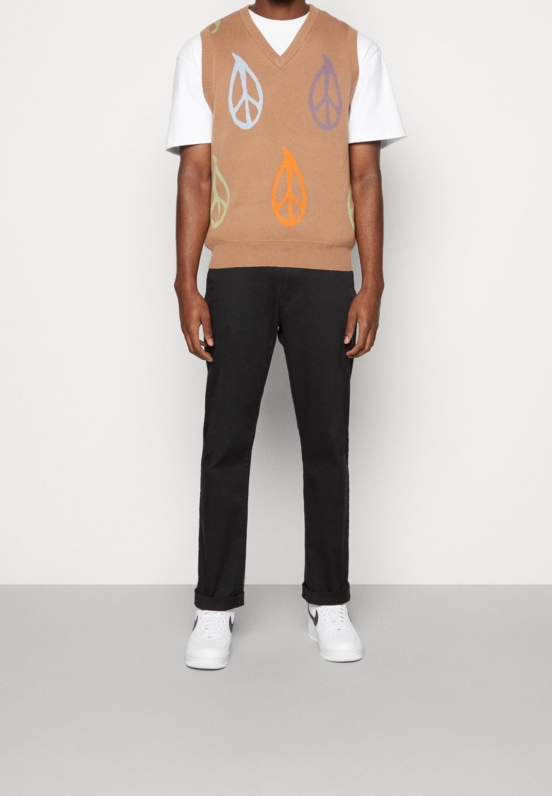 Beiger Strickwest mit V-Ausschnitt, verziert mit bunten Blattmustern in Blau, Orange, Lila und Grün, getragen über einem weißen T-Shirt und schwarzen Hosen.