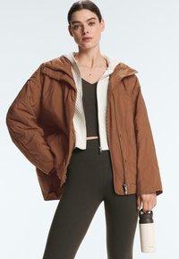 Kvinde iført en brun quiltet jakke over en hvid lynlås-sweater og sorte leggings, der holder en beige isoleret vandflaske i sin højre hånd.