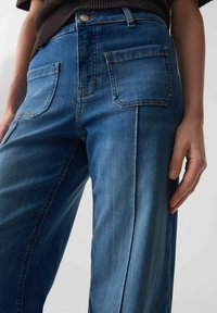 Hoch taillierte blaue Denim-Jeans mit lockerer Passform, sichtbaren Nähten und zwei Gesäßtaschen. Der Stoff hat eine glatte Textur und einen leichten Glanz.