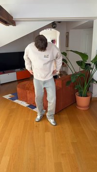 Sweatshirt cinza claro com logótipo, jeans azul claro e ténis prateados metálicos. Em pé numa sala com um sofá castanho e uma planta verde.