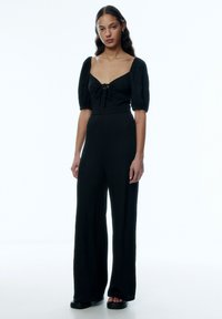 Schwarzer Jumpsuit mit Puffärmeln und quadratischem Ausschnitt. Strukturiertes Gewebe, Kordelzugdetails vorne, weite Beine und flache Sandalen.
