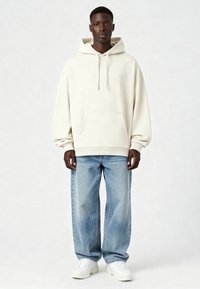 Mikina s kapucňou krémovej farby s klokankovým vreckom, doplnená o svetlomodré široké rifle a biele tenisky. Mäkká textúra, pohodlný strih.