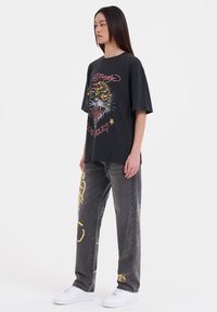 Černé oversized tričko s barevným grafikou tygra, doplněné šedými džíny s žlutým textem a akcenty, dokončeno bílými teniskami.