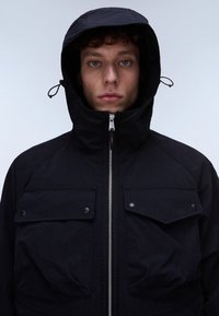 Ung mand med krøllet hår iført en sort hættetrøje med lynlås, vendt mod fronten mod en ensartet lys baggrund.