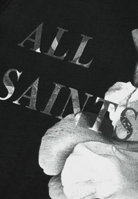 Juodas marškinėliai su dideliu grafiniu spausdinimu, vaizduojančiu rankas ir gėles, su tekstu „ALL SAINTS“ drąsiais, nusidėvėjusiais sidabro spalvos šriftais.
