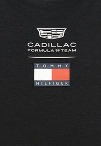 Λογότυπο της ομάδας Formula 1 της Cadillac πάνω από το ορθογώνιο σχέδιο σημαίας της Tommy Hilfiger σε λευκό, κόκκινο και μπλε, σε μαύρο φόντο υφάσματος.