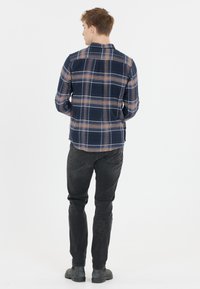 Marineblauw geruit overhemd van zachte stof, met bruine en beige lijnen. Gepaard met zwarte jeans en grijze schoenen, gezien vanuit de achterkant.