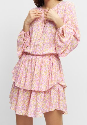 Femme portant une robe à manches longues à motifs floraux roses et jaunes avec une jupe à volants à étages, ajustant les liens du décolleté.