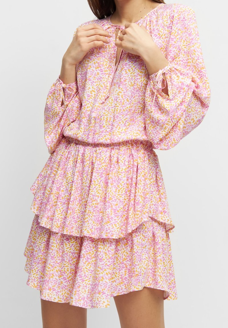 Femme portant une robe à manches longues à motifs floraux roses et jaunes avec une jupe à volants à étages, ajustant les liens du décolleté.