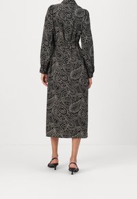 Czarna sukienka w paisley z długimi rękawami, paskiem w talii i długości midi; wykonana z gładkiego, zwiewnego materiału. Wyeksponowane czarne sandały na obcasie.