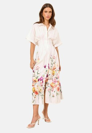 Femme portant une robe midi blanche à manches courtes avec un imprimé floral coloré le long de la jupe, associée à des sandales blanches à talons hauts.