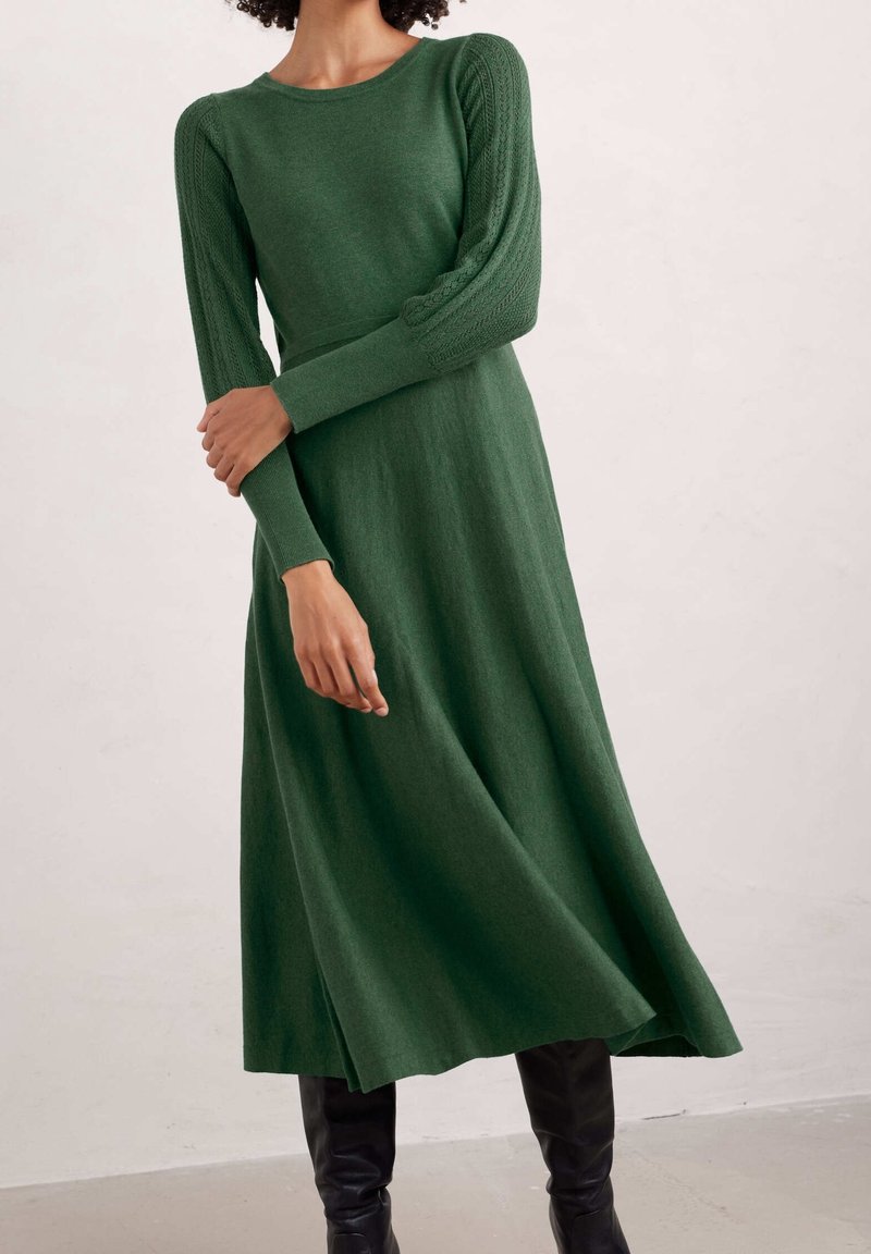 Femme portant une robe midi en tricot vert à manches longues, associée à des bottes noires montantes jusqu'aux genoux, debout contre un mur uni.