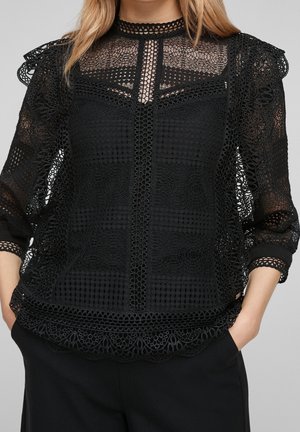 Blouse en dentelle noire avec des motifs complexes, un col montant et des bords festonnés, portée avec un pantalon noir et les mains dans les poches.