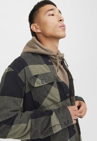 Camisa de botones en un patrón de cuadros verde y negro. Tela suave y texturada; dos bolsillos en el pecho; llevada sobre una sudadera con capucha marrón.