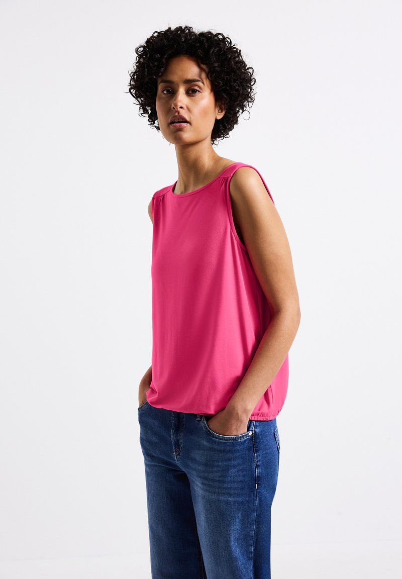 Top senza maniche di colore rosa realizzato in tessuto morbido, con scollatura rotonda e leggeri volant sulle spalle, abbinato a jeans blu.