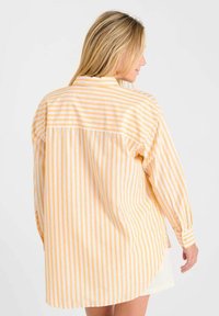 Femme aux cheveux blonds portant une chemise ample à manches longues avec des rayures verticales et horizontales jaunes et blanches, de dos contre un fond uni.