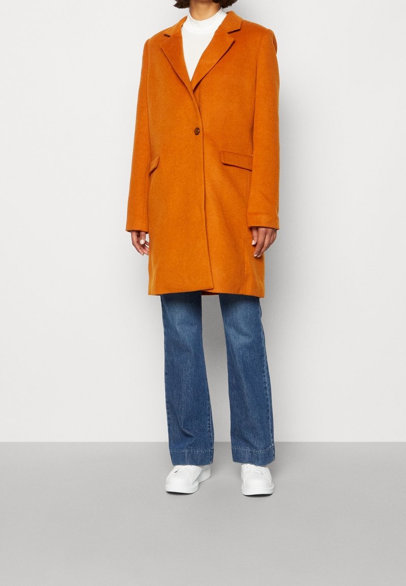 Abrigo de mezcla de lana naranja con solapa de muesca, cierre de un solo botón, dos bolsillos frontales y corte relajado, combinado con jeans azul de campana y zapatos blancos.